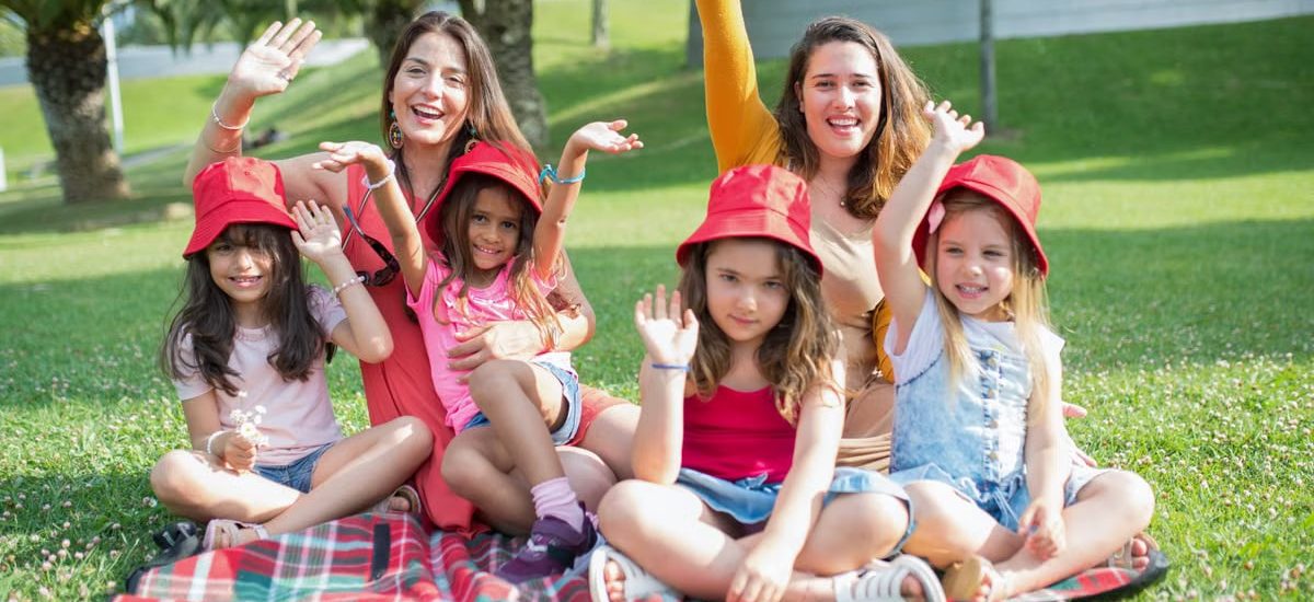 Two women and four girls enjoy a sunny picnic in Portugal, waving happily in the park.