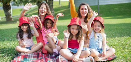 Two women and four girls enjoy a sunny picnic in Portugal, waving happily in the park.
