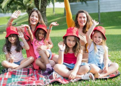 Two women and four girls enjoy a sunny picnic in Portugal, waving happily in the park.